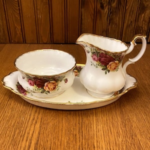 Puede incluir: Un juego de azucarero y jarra de crema de porcelana blanca con un estampado floral de rosas y ribetes dorados. El juego incluye una jarrita de crema, un azucarero y una bandeja ovalada a juego.
