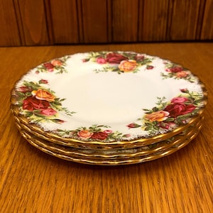 Puede incluir: Un juego de cuatro platos pequeños de cerámica blanca con borde dorado y un patrón floral con rosas rojas, rosas y amarillas. Los platos están apilados uno encima del otro.