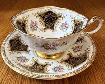 May include: A vintage teacup and saucer set. The white porcelain cup and saucer feature a black and gold ornate design with floral accents. The cup has a gold handle and sits on a gold base. The set is displayed on a wooden surface.