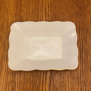 Puede incluir: Un plato pequeño, rectangular, blanco con un borde dorado. El plato tiene un borde ligeramente festoneado y está hecho de porcelana.
