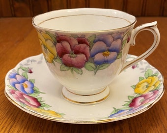 May include: A vintage teacup and saucer set with a floral pattern featuring colorful pansies. The set is white with a gold trim.
