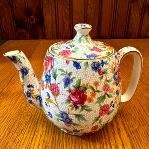 May include: A white teapot with a floral pattern in shades of pink, blue, yellow, and green. The teapot has a gold trim around the lid and spout.