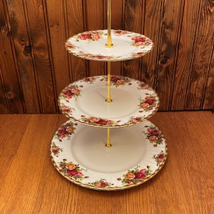 Puede incluir: Un soporte para tartas de tres niveles con un marco de metal dorado y platos de porcelana blanca decorados con un patrón floral de rosas rojas, rosas y amarillas.