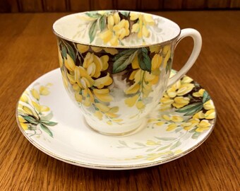 May include: A white teacup and saucer with a floral pattern. The cup and saucer are decorated with yellow flowers and green leaves. The cup has a gold rim.