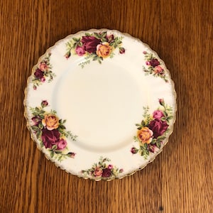 Puede incluir: Un plato de cerámica blanco con un borde dorado y un diseño floral con rosas rojas, rosas y amarillas. El plato tiene un borde festoneado.
