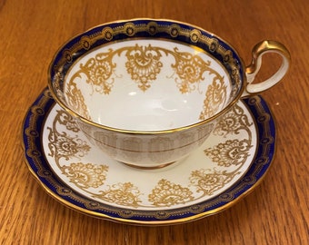 May include: A white teacup and saucer set with a gold and navy blue floral design. The teacup has a gold handle and a navy blue rim. The saucer has a matching navy blue and gold border.