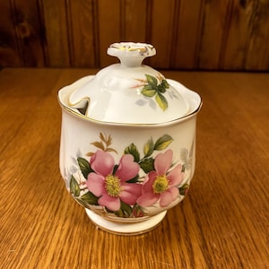 Puede incluir: Un azucarero de cerámica blanca con tapa, con un diseño floral con rosas rosas y hojas verdes. El azucarero tiene un borde dorado alrededor del borde y la base.