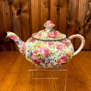 Puede incluir: Una tetera con estampado floral y tapa a juego. La tetera presenta una base blanca adornada con rosas rosas, flores azules y amarillas y hojas verdes. El asa y el pico son curvos, y la tapa tiene un pequeño pomo. La tetera se exhibe sobre un soporte acrílico transparente.