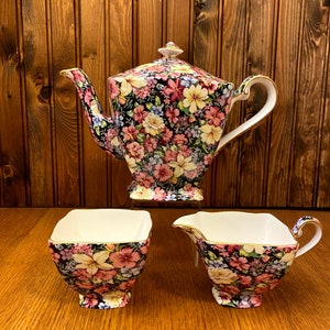 May include: A floral tea set comprising a teapot, creamer and sugar bowl. The set features a black background with a colourful floral pattern.