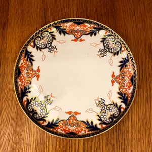 May include: A white ceramic plate with a blue and gold rim and a red and gold floral design.
