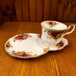 Puede incluir: Un juego de taza de té, platillo y plato de servir blancos con un estampado floral de rosas en rojo, rosa y amarillo. Los bordes están adornados con dorado. El juego está sobre una superficie de madera.