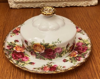 May include: A white ceramic butter dish with a floral rose pattern in red, yellow, and pink. The dish has a gold-colored rim and a decorative gold knob on the lid. The dish sits on a wooden surface.
