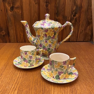 Könnte beinhalten: Ein Vintage-Blumenservice mit einer Teekanne und zwei Tassen und Untertassen. Die Teekanne und die Tassen sind mit einem gelben, violetten und grünen Blumenmuster verziert.
