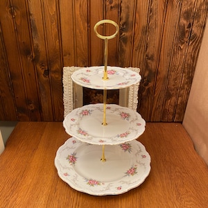 Könnte beinhalten: Ein dreistöckiger Kuchenständer mit einem goldenen Metallständer und weißen Porzellanplatten mit einem Blumendesign. Die Teller haben ein rosa-weißes Blumendesign mit goldenen Verzierungen.