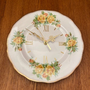 May include: A white porcelain plate with a gold rim and a floral design featuring yellow roses. The plate has been converted into a clock with Roman numeral markings and gold hands.