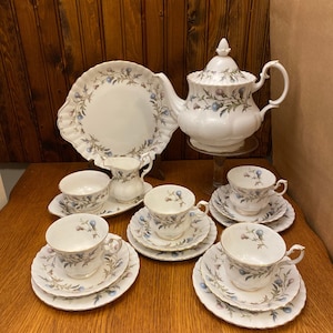 Peut inclure: Service à thé vintage blanc orné d'un motif floral de baies bleues et violettes et de feuilles vertes. L'ensemble comprend une théière, une assiette de service, une crémière, un sucrier et six tasses à thé avec soucoupes. Les bords sont dorés.