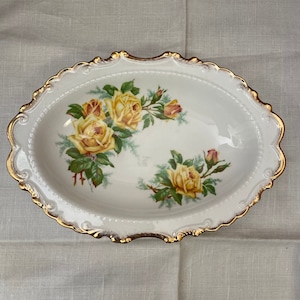 Könnte beinhalten: Eine weiße ovale Servierplatte mit goldfarbenem Rand und einem Blumendesign mit gelben Rosen. Die Rosen sind in einem realistischen Stil mit grünen Blättern und Stielen gemalt.