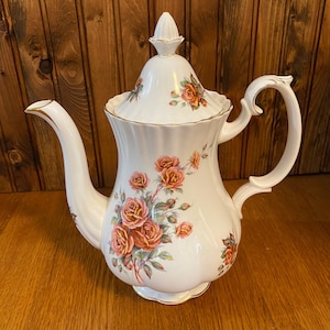 Może przedstawiać: Biały porcelanowy czajnik z kwiatowym wzorem przedstawiającym różowe i pomarańczowe róże. Czajnik ma złotą obwódkę wokół pokrywki i podstawy.