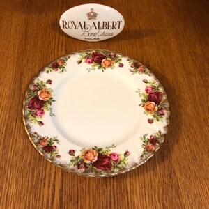 Puede incluir: Un plato de porcelana blanca con un borde dorado y un patrón floral con rosas rojas, rosas y amarillas. El plato está hecho por Royal Albert Bone China, Inglaterra.