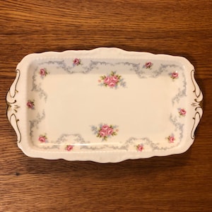 Könnte beinhalten: Ein rechteckiges Serviertablett in Weiß mit goldfarbenem Rand und einem zarten Blumenmuster in Rosa- und Blautönen.