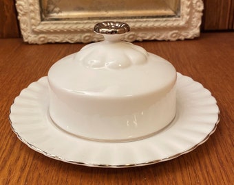 May include: White ceramic butter dish with a silver-trimmed lid and a scalloped edge. The dish is sitting on a wooden surface.