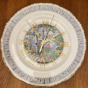 Peut inclure: Une assiette en porcelaine blanche avec un motif floral bleu et vert et un cadran d'horloge en chiffres romains. L'assiette a un bord doré et un motif d'écorce de bouleau gris et blanc autour du bord.