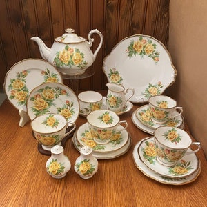 May include: A complete vintage tea set, including a teapot, plates, cups, saucers, a creamer, and salt and pepper shakers. The white porcelain is decorated with yellow rose patterns and gold edging, displayed on a wooden table.