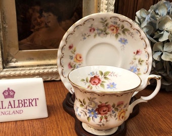 May include: A white teacup and saucer with a floral pattern. The teacup has a gold trim and the saucer has a scalloped edge. The teacup and saucer are sitting on a wooden surface. The text "ROYAL ALBERT ENGLAND" is visible on a white rectangular sign.