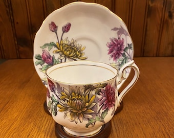 May include: A white teacup and saucer with a floral pattern featuring pink, yellow, and green chrysanthemums. The cup and saucer have a gold trim.