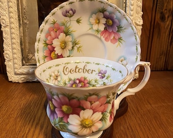 May include: A white teacup and saucer with a floral design. The teacup has the word "October" written on it in gold lettering. The saucer has a delicate floral pattern with pink, purple, and white flowers.