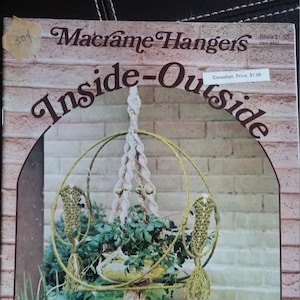 May include: A book cover with the title "Macrame Hangers Inside-Outside" featuring a macrame plant hanger with a white cord and a green plant inside. The book cover is brown with a brick wall background.