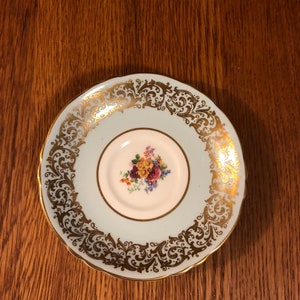 May include: A light blue saucer with a gold floral design around the edge. The center of the saucer has a white floral design.