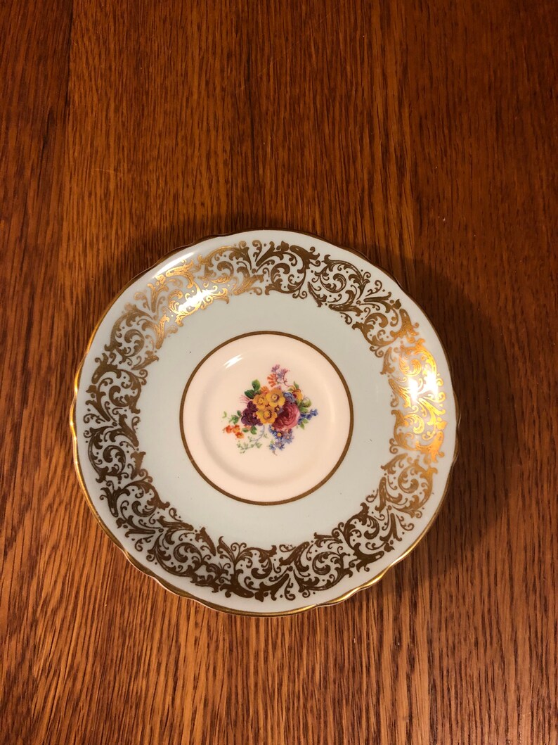 May include: A light blue saucer with a gold floral design around the edge. The center of the saucer has a white floral design.