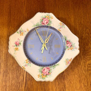 May include: A vintage wall clock with a light blue face and gold hands and numerals. The clock is surrounded by a cream-colored border decorated with pink roses and green leaves. The clock is set on a wooden surface.