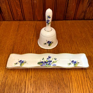 Puede incluir: Una campana de porcelana blanca y una bandeja rectangular con borde ondulado. Ambas piezas están decoradas con diseños florales azules y morados y ribetes dorados. La campana tiene un asa con detalles florales a juego.