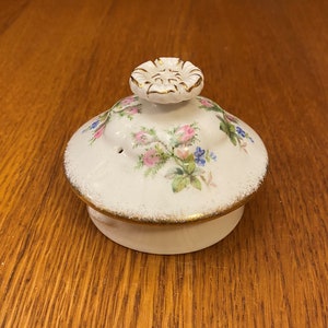 Könnte beinhalten: Weißer Teekannen-Deckel aus Keramik mit einem Blumendesign in Rosa, Blau und Grün. Der Deckel hat einen goldenen Rand und einen kleinen Knopf in der Mitte.