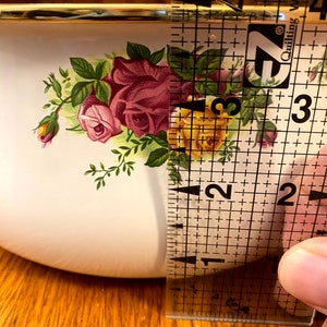 May include: A white ceramic bowl with a gold rim and a floral pattern featuring pink and yellow roses. The bowl is being measured with a ruler, showing it is approximately 2 inches in diameter.