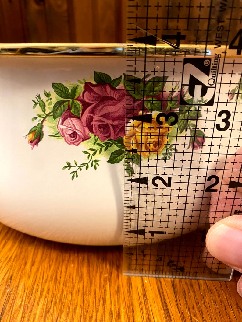 May include: A white ceramic bowl with a gold rim and a floral pattern featuring pink and yellow roses. The bowl is being measured with a ruler, showing it is approximately 2 inches in diameter.