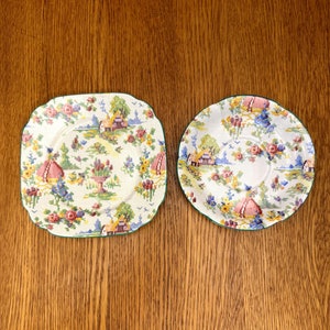 Puede incluir: Dos platos de cerámica blanca con un borde verde y un patrón floral. Los platos presentan un diseño cottagecore con una pequeña casa, flores y pájaros.