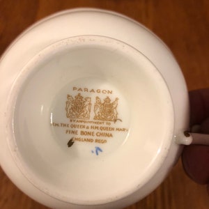 May include: Close-up of the bottom of a white teacup with a gold logo that reads "PARAGON BY APPOINTMENT TO H.M. THE QUEEN & H.M. QUEEN MARY FINE BONE CHINA ENGLAND REGO."