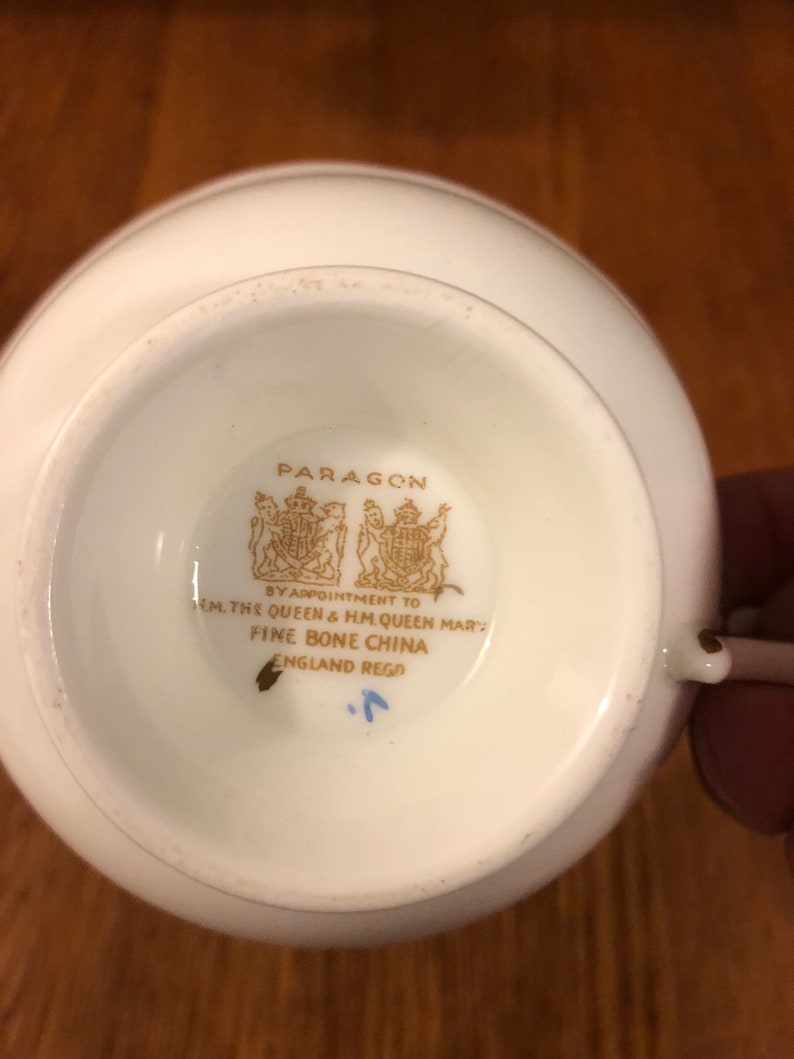 May include: Close-up of the bottom of a white teacup with a gold logo that reads "PARAGON BY APPOINTMENT TO H.M. THE QUEEN & H.M. QUEEN MARY FINE BONE CHINA ENGLAND REGO."