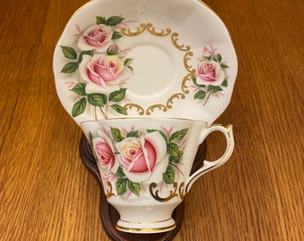 Könnte beinhalten: Eine weiße Teetasse und Untertasse mit einem rosafarbenen Rosenblütenmuster und goldenen Akzenten. Die Teetasse und Untertasse befinden sich auf einer Holzoberfläche.