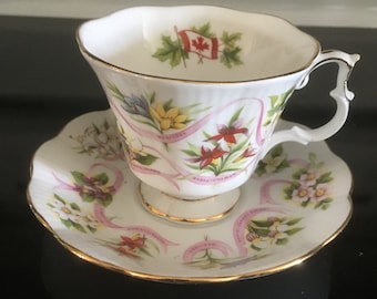 May include: White teacup and saucer with a gold rim. The cup and saucer are decorated with pink ribbons and floral designs representing the provinces of Canada. The cup has a Canadian flag on the inside.