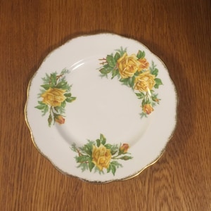 May include: A white porcelain plate with a gold rim features a floral design of yellow roses with green leaves.