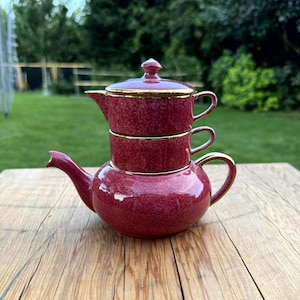 Puede incluir: Juego de té burdeos apilado con ribete dorado, que incluye una tetera, dos tazas y una tapa. El juego está colocado sobre una mesa de madera con un fondo verde. La tetera tiene un pico y un asa curvos.
