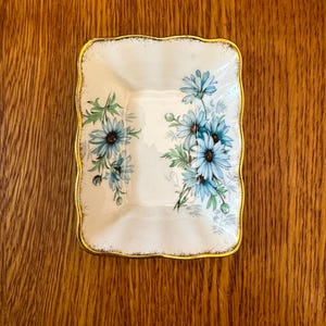 May include: A square, off-white dish with a gold trim and a floral design. The flowers are blue with green leaves. The dish is sitting on a wooden surface.