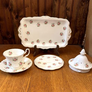 Könnte beinhalten: Ein weißes Porzellan-Teeservice mit rosa Rosenmuster. Das Set beinhaltet eine Teetasse und Untertasse, ein Serviertablett, einen kleinen Teller und einen Deckel. Die Ränder sind mit Gold verziert. Das Teeservice wird auf einer Holzoberfläche präsentiert.