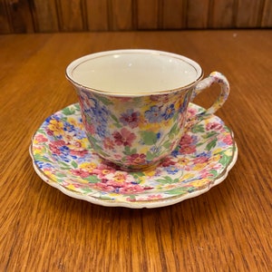 Puede incluir: Un juego de taza y platillo con estampado floral. La taza y el platillo están decorados con un patrón de pequeñas flores coloridas en tonos rosa, azul, amarillo y verde sobre un fondo blanco. Los bordes están adornados con dorado. El juego está sobre una superficie de madera.