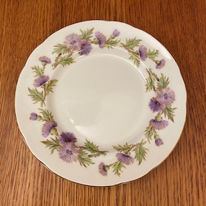 Könnte beinhalten: Weißer Keramikteller mit gewelltem Rand und Goldrand. Der Teller ist mit einem floralen Muster aus lila Disteln und grünen Blättern verziert. Auf einer Holzoberfläche platziert.