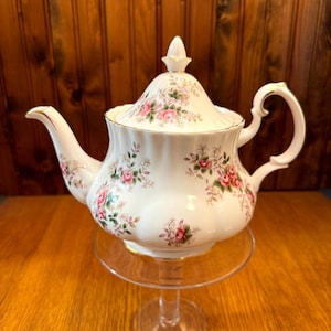 Op de afbeelding: Een witte theepot met een bloemenpatroon in roze en groene tinten. De theepot heeft een gouden rand rond de deksel en de basis. Hij staat op een glazen voet.
